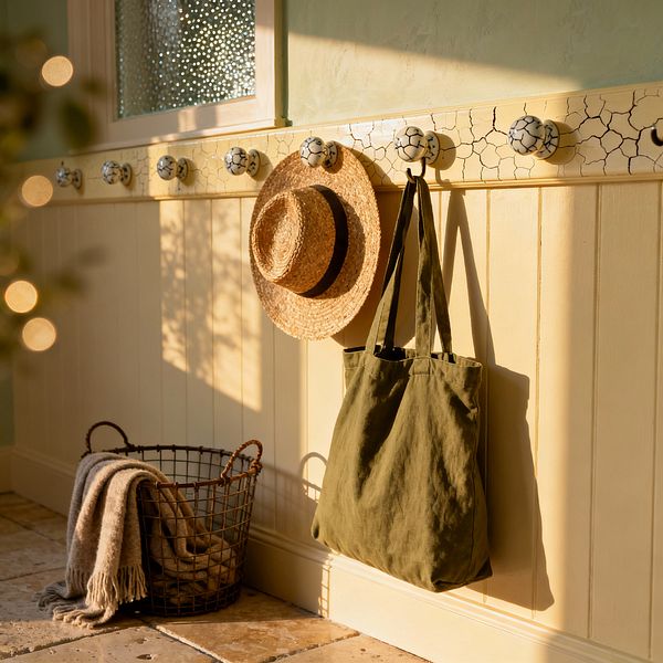 A sunlit mudroom features cream tongue-and-groove paneling topped by a crackle-finish peg rail with round ceramic hooks. An olive canvas tote and a straw hat hang from the hooks, while a metal wire basket holding a beige fringed blanket sits on tumbled limestone tiles; the upper wall is painted pale sage and a small window with textured glass diffuses the warm light.