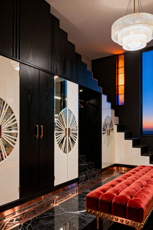 A luxurious Art Deco mudroom features black, stair-stepped cabinetry paired with glossy ivory doors inset with mirrored sunburst motifs. A polished black marble floor with copper inlay borders a tufted red velvet bench with brass studs. A tiered alabaster pendant hangs above, while a vertical stained-glass panel in amber and cobalt glows beside black stone steps, with evening sky visible through a tall window.