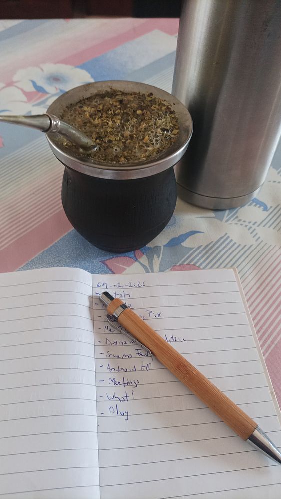 A person sitting outdoors on a raised concrete ledge beside a glass railing, enjoying the sunny weather with a beverage in hand and looking thoughtful.