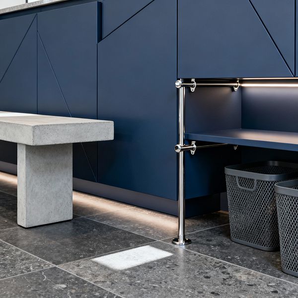 A modern mudroom features flat-front navy cabinets with subtle geometric grooves, a built-in navy bench supported by polished chrome tubing, and integrated LED strip lighting at the shelf and toe kick. The floor is laid in large-format honed gray stone tiles, a solid concrete bench sits opposite, and two gray open-weave plastic baskets are tucked neatly beneath the bench for storage.