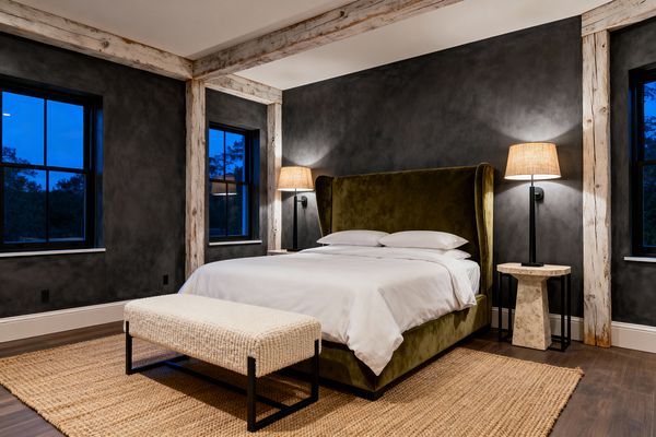 A moody transitional bedroom features troweled charcoal plaster walls framed by rustic whitewashed beams and black-trimmed windows. At center sits an olive green velvet wingback bed dressed in white linens, flanked by stone pedestal nightstands and black metal wall sconces with woven shades; a large jute rug covers dark wood flooring, and a cream textured bench with black metal legs rests at the foot of the bed.