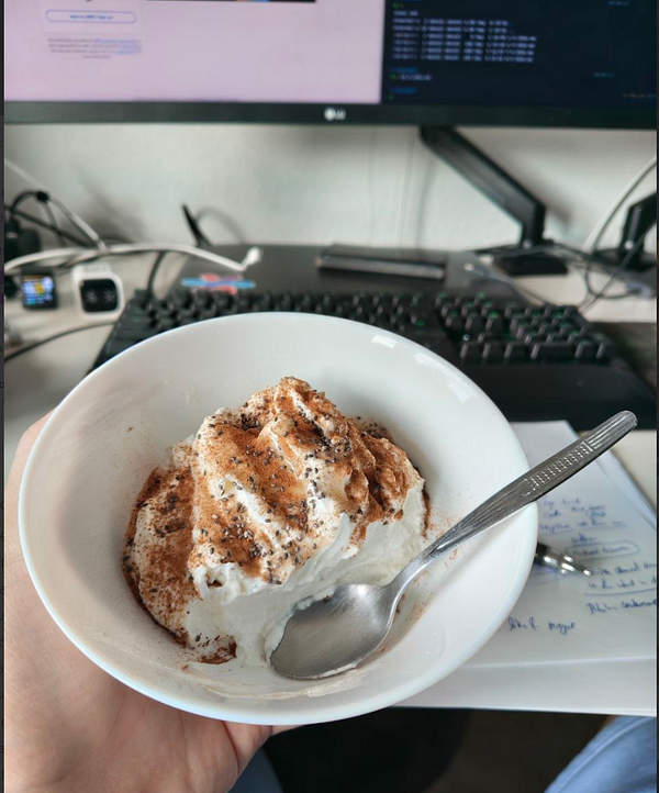 A transparent cup containing white yogurt topped with chia seeds, honey, and a dusting of cinnamon is placed on a table with other breakfast items.