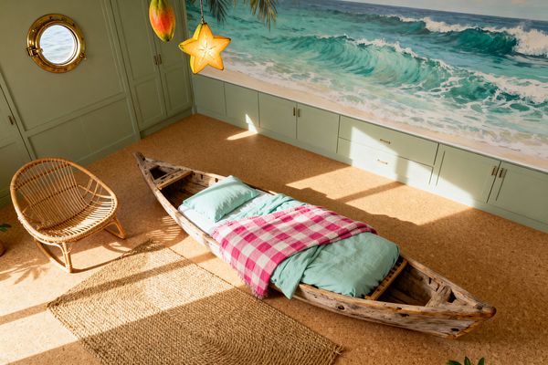 Sunlit kids’ room with a weathered wooden canoe used as a bed, dressed in aqua bedding and a pink-and-white gingham throw. A curved rattan chair and woven jute rug sit on a sand-toned cork floor. Mint green built-in cabinets with brass pulls run beneath a large beach-and-waves mural, while a round brass porthole window and hanging fruit-shaped pendants complete the tropical, nautical theme.