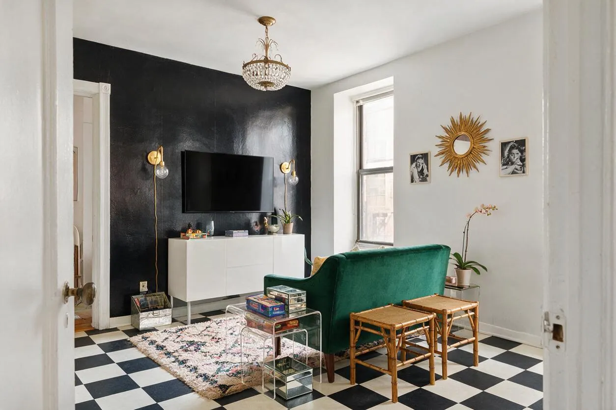 A compact game room with black-and-white checkerboard tile flooring, a glossy black accent wall, and a wall-mounted TV above a white credenza. A deep green velvet sofa, clear acrylic side tables, bamboo stools, and gold accents create a glamorous Hollywood Regency look, with a crystal chandelier and sunburst mirror adding sparkle.