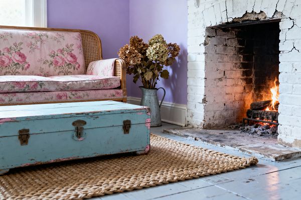 A shabby chic living room features a pink floral-upholstered rattan sofa against lavender walls beside a white-painted brick fireplace with a live fire. A distressed turquoise trunk with pink accents serves as a coffee table on a chunky jute rug over pale gray-blue floorboards, while a galvanized metal pitcher filled with dried hydrangeas sits near the hearth and daylight from a window with white trim softens the room’s pastel palette and worn textures.