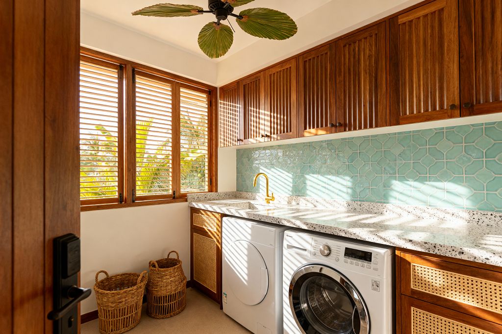 A bright tropical laundry room features wooden louvered windows casting striped sunlight across a terrazzo countertop above a white front-loading washer and dryer. Slatted wood upper cabinets pair with cane-front base cabinets. A brushed brass faucet sits at an undermount sink before a glossy seafoam arabesque tile backsplash. Two rattan baskets rest on a sandy floor, and a ceiling fan with palm-leaf blades hangs overhead.
