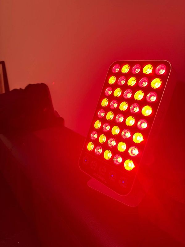 Hand holding a handheld red light therapy device emitting red light, targeting the thyroid area on the neck.