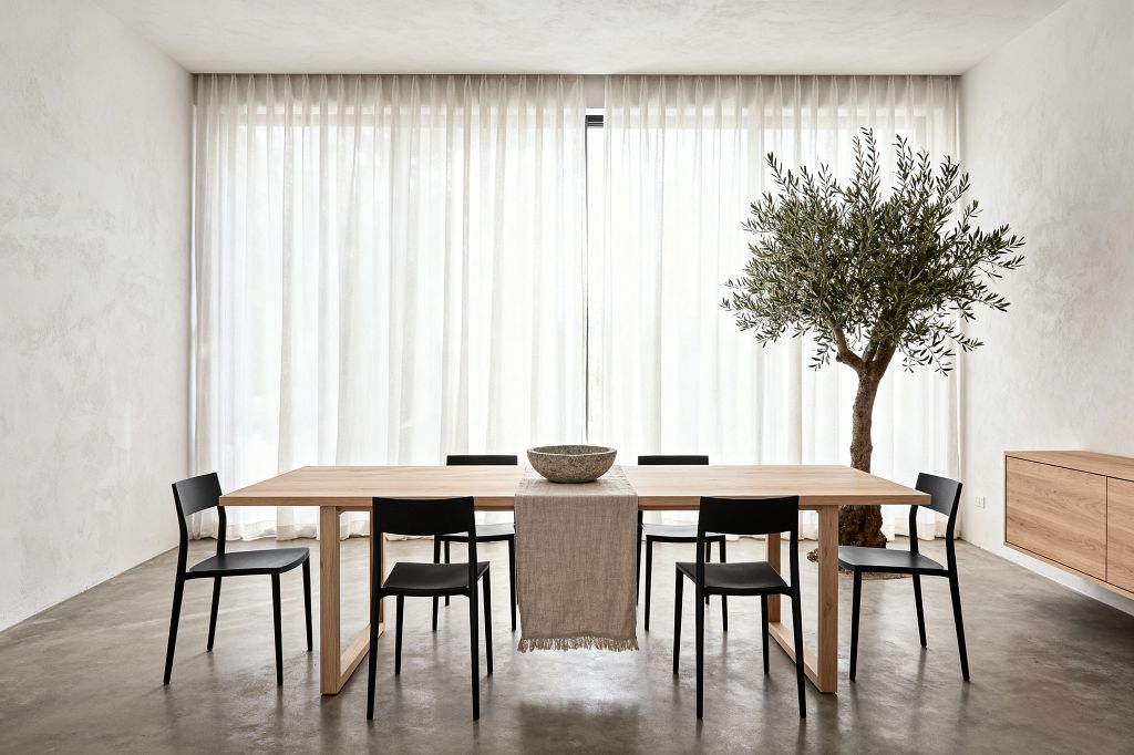 A minimalist dining room with a long light oak table centered on a polished concrete floor, surrounded by six matte black chairs. Floor-to-ceiling sheer white curtains diffuse daylight across warm white plaster walls. A neutral linen runner and a stone bowl sit on the tabletop. An indoor olive tree softens the right side beside a wall-mounted oak sideboard. The space feels uncluttered, airy, and calm with clean lines and natural textures.