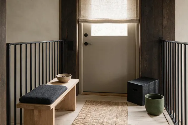 A narrow front porch styled in Japandi design with a light wood bench, dark cushion, woven jute runner, and simple ceramic bowls and planters. The space features a beige front door, dark wood wall panels, black metal railings, and a soft linen shade that filters daylight.