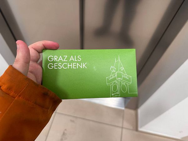 A hand holds a green voucher for Graz, Austria, in front of an elevator.