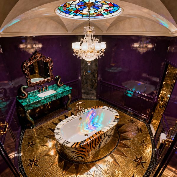 An opulent bathroom centers on a freestanding white tub with a tiger-striped, marble-clad exterior, set on a gold mosaic floor with a starburst motif. Glossy purple paneled walls frame a green malachite-style vanity with sculptural serpent legs and an ornate mirror, while brass faucets and a crystal chandelier gleam beneath a domed ceiling with a round stained-glass skylight casting multicolored reflections; a doorway reveals a dark marble shower.
