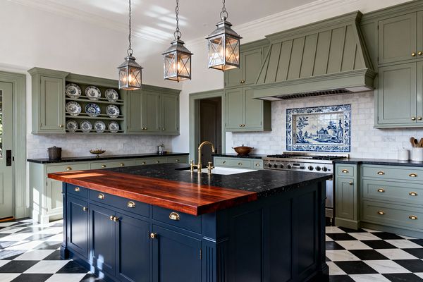 A traditional kitchen features sage green Shaker-style cabinets and a navy blue island finished with honed black stone and a thick wood butcher-block end, accented by brass bin pulls and a brass bridge faucet. A marble subway backsplash with a blue-and-white Delft mural sits behind a stainless range, three lantern pendants illuminate the island, a black-and-white checkerboard marble floor grounds the space, and open plate racks display blue-and-white china.