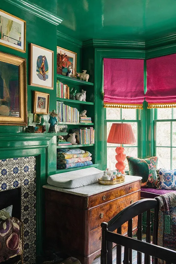 A maximalist nursery with glossy emerald-green walls, built-in shelves filled with children’s books, framed artwork, and small decorative animals. The room features magenta Roman shades, a coral lamp, a vintage wood changing dresser with a pale changing pad, patterned pillows, and a black crib in the foreground.