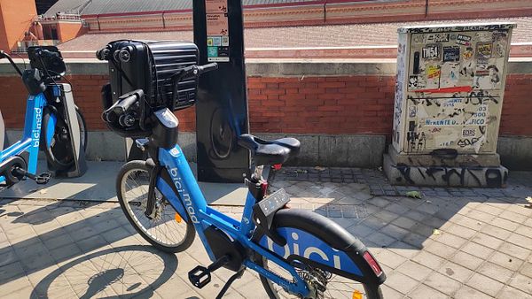 A blue bike with a suitcase attached is parked at a bike station.