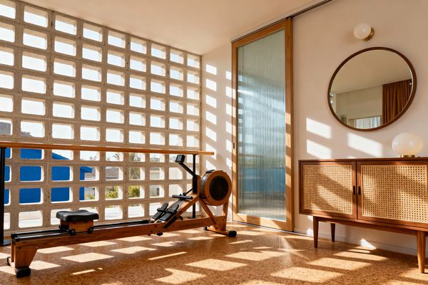A bright home gym with a wooden rowing machine and a wall-mounted barre faces a concrete breeze-block screen that filters light into grid-like shadows across a cork floor. At right, a ribbed-glass sliding door in a wood frame sits beside a walnut credenza with cane-front doors, a round wood-framed mirror, and globe lighting, all in warm white and honey wood tones with subtle black metal accents.