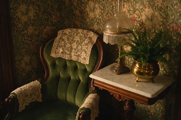 A cozy Victorian-style corner features a deep green velvet armchair with carved wood trim, decorated with delicate lace doilies on the back and arms. Beside it sits a small marble-topped table with a brass lamp and a gold planter, set against richly patterned floral wallpaper in warm, dim light.