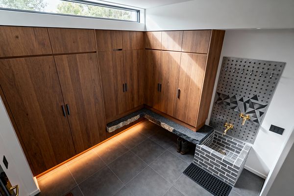 A contemporary mudroom features full-height L-shaped walnut cabinets with black pulls and warm LED lighting at the toe-kick. A floating dark stone bench with a chiseled edge lines the corner, while a gray brick dog-wash basin sits below a perforated gray tile splashback with a geometric band, brass wall-mounted taps, and metal hooks; large-format gray porcelain floor tiles and a narrow clerestory window complete the clean, durable space.