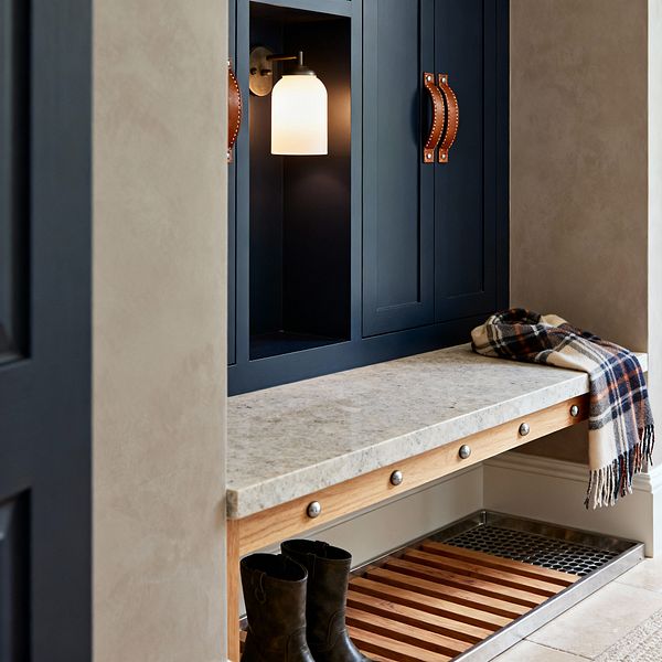 Close-up of a transitional mudroom built-in with deep navy Shaker cabinets and cognac leather strap handles. A light, speckled stone bench spans the unit, with an opal-glass brass sconce glowing inside a recessed cubby; below, a natural wood slatted shelf sits above a stainless boot tray, while warm beige walls and stone-look floor tiles complete the scene, with a plaid scarf on the bench and boots tucked underneath.