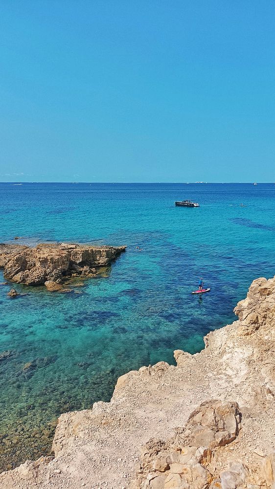 A scenic view of a clear blue ocean with rocky shores and a paddleboarder.