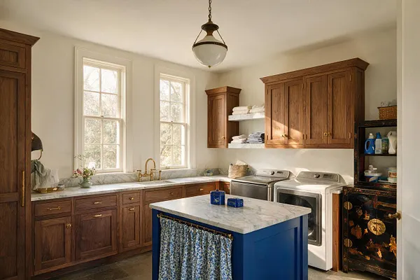 A bright eclectic laundry room with warm wood cabinets, white walls, and two tall windows bringing in natural light. A blue island with a marble top sits in the center, while a black lacquered cabinet with gold decoration adds a bold artistic accent beside the washer and dryer.