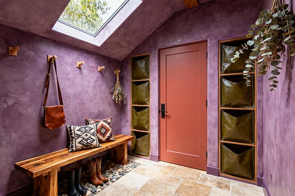 A compact mudroom features textured plum-lavender walls and a pitched ceiling illuminated by a large skylight. A live-edge wood bench with kilim pillows sits over a recessed river-rock boot tray, oak pegs hold a leather bag and dried branches, olive-green leather sling cubbies line the wall, and a terracotta-coral door with matte-black hardware opens onto tumbled beige stone flooring.