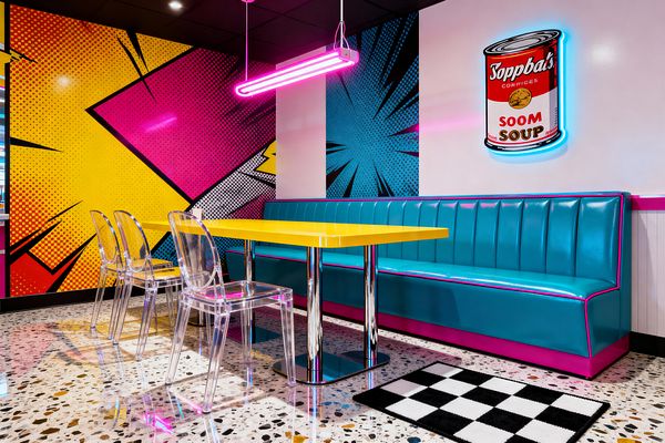 A Pop Art dining nook with a long teal vinyl banquette trimmed in hot pink faces a glossy yellow rectangular table on chrome pedestal bases. Three clear polycarbonate chairs sit on speckled terrazzo flooring beside a small black-and-white checkerboard rug. A bold comic-style mural in yellow, magenta, and cyan covers the walls, while a pink neon pendant and a neon-outlined soup can artwork glow beneath a black grid ceiling.