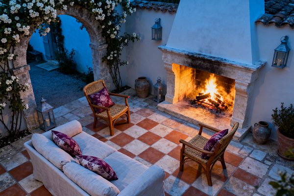 At dusk, a French Country patio features a blazing outdoor fireplace with a carved limestone surround and white plaster chimney. A linen slipcovered sofa and two rattan armchairs with plum floral pillows sit on a terracotta-and-limestone checkerboard floor, while galvanized lanterns, clay urns, and a weathered stone arch draped in white climbing roses frame the space against whitewashed walls and rustic roof tiles.