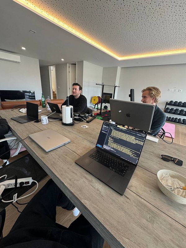 A modern coworking space with three individuals working at a large wooden table, surrounded by laptops and fitness equipment.