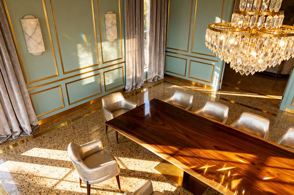 A sunlit dining room with pale sage paneled walls trimmed in gold, a large tiered crystal chandelier, and a polished walnut rectangular table on a brass plinth base. Champagne-upholstered armchairs with wooden legs surround the table, gray velvet floor-length curtains frame tall windows, terrazzo flooring with brass inlays gleams, and white marble wall sconces add sculptural accents for a vintage glam ambiance.