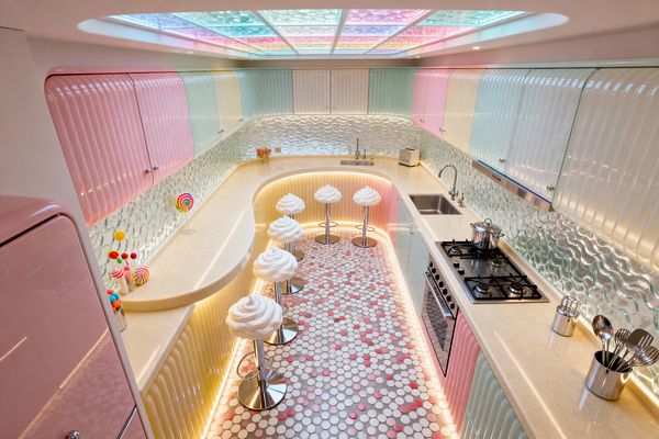 A narrow U-shaped kitchen styled like Candy Land features glossy ribbed cabinets in pastel pink, mint, and butter yellow, cream solid-surface countertops, and a wavy textured glass backsplash. A curved breakfast bar seats five on white swirl-topped stools with chrome bases. The ceiling glows through multicolor backlit panels, while warm LED strips outline counters and toe-kicks. The floor is a penny tile mosaic in white, gray, and pink with lollipop decor on the counter.