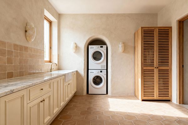 A transitional laundry room features a stacked white washer and dryer tucked into an arched plaster niche, framed by two alabaster wall sconces. On the left, cream raised-panel cabinets with brass knobs hold a gray‑veined marble countertop and a white apron-front sink beneath a wood-framed window. A square tan zellige tile backsplash lines the wall, terracotta hex tiles cover the floor, and a tall slatted oak storage cabinet stands to the right.