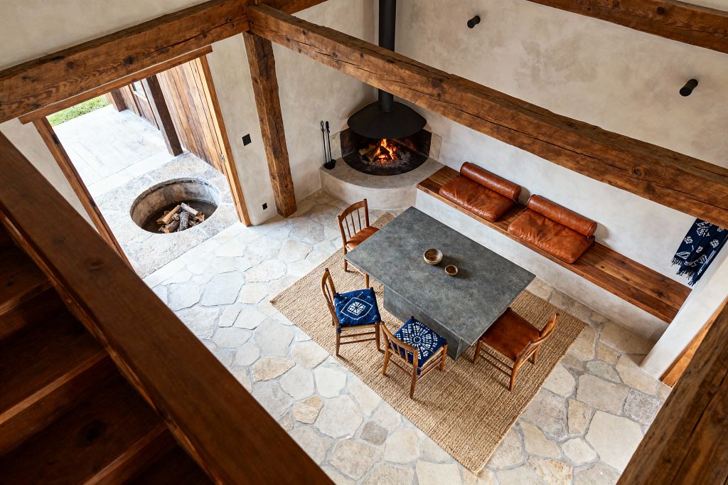 A rustic living room with exposed timber beams, pale plaster walls, and irregular flagstone flooring. A black suspended corner fireplace sits above a curved stone hearth beside a built-in wooden bench topped with caramel leather cushions; at center, a gray concrete table on a jute rug is surrounded by simple wooden chairs, two with indigo patterned cushions, while daylight enters through an open wood door to a patio with a circular stone fire pit.