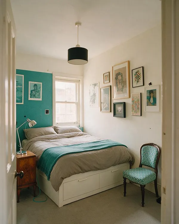 A cozy guest bedroom with a bed centered under a window, dressed in gray bedding and a teal throw, with a bright teal accent wall at the headboard. The room includes a wooden nightstand, a chrome reading lamp, a patterned teal side chair, and a gallery wall of mixed framed artwork on the white wall.