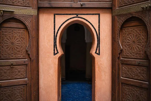 A Moroccan-style exterior entrance features richly carved wooden doors on both sides of a peach-toned stucco wall. A central horseshoe arch outlined in dark tile opens into a dim interior with a blue patterned floor.