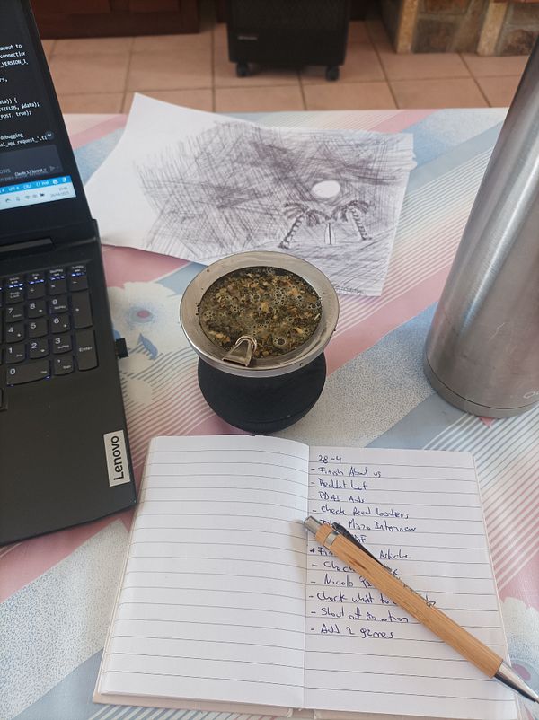 A workspace featuring a laptop, a notebook with a to-do list, a traditional mate drink, and a sketch in the background.
