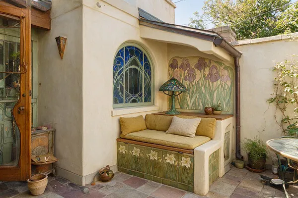 A small enclosed patio features an Art Nouveau-style built-in bench with floral tile panels, mustard cushions, and a stained-glass window above. A hand-painted iris mural, curved stucco walls, and a decorative mosaic table create a soft, romantic courtyard atmosphere.