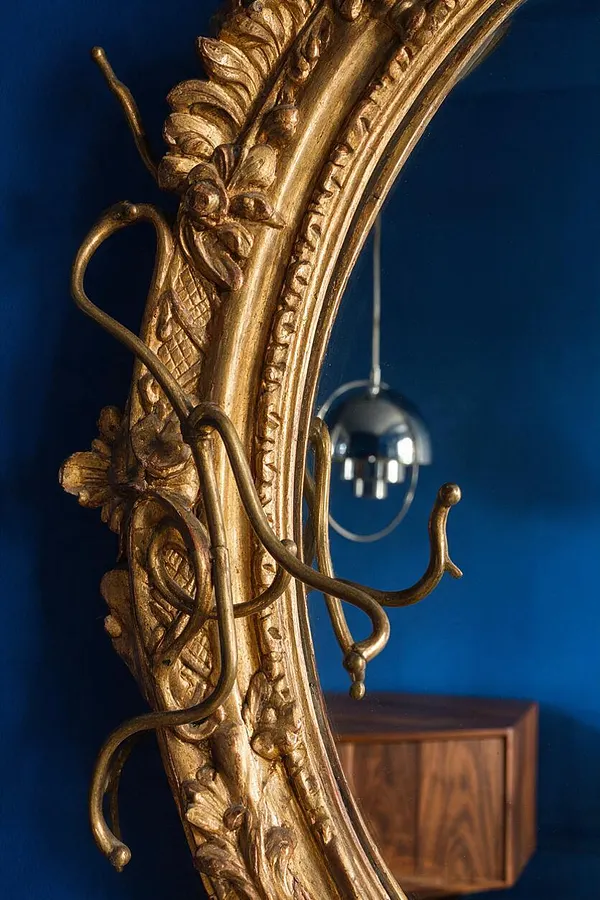 A close-up of an ornate gilded mirror frame with carved floral details and curling metal hooks in front of a deep cobalt blue wall. A modern chrome pendant light and a walnut cabinet appear softly in the background, creating a layered eclectic contrast.