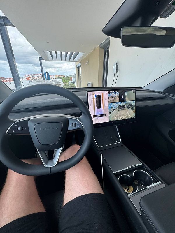 The image captures the interior of a Tesla vehicle, showcasing the steering wheel, touchscreen display, and the driver's legs.