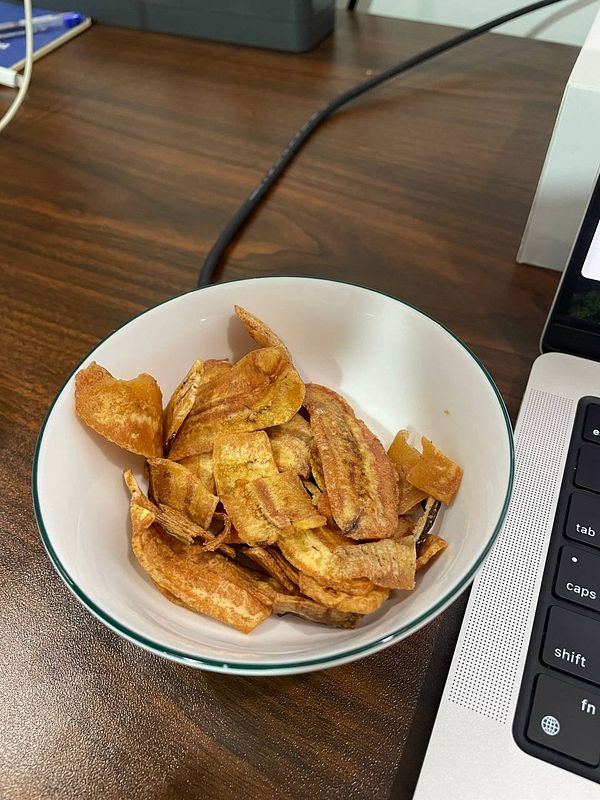 A bowl of dried banana snacks is placed on a wooden table next to a laptop.