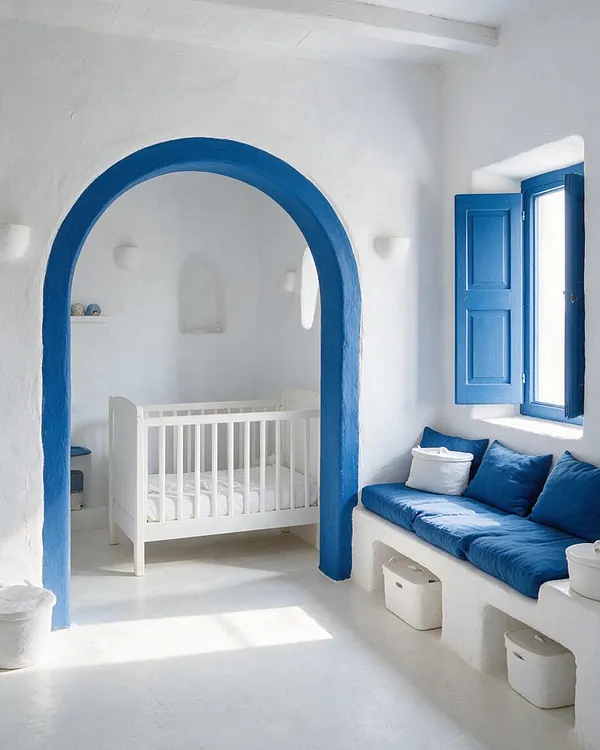 A bright Mediterranean-style nursery with white plaster walls, a vivid blue arched doorway, and matching blue window shutters. A white crib sits in the adjoining alcove, while a built-in bench with deep blue cushions lines the right wall under the window.