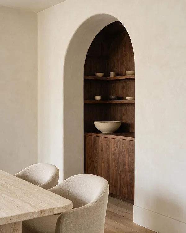 A serene dining room in an organic modern style with a rounded plaster arch framing a walnut built-in niche. Open wooden shelves display simple ceramic bowls, while a pale stone dining table and curved upholstered chairs sit in the foreground against warm neutral walls and light wood flooring.