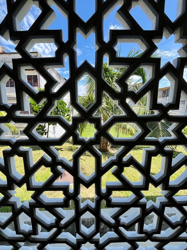 A decorative lattice window frame offers a view of a lush garden and blue sky.