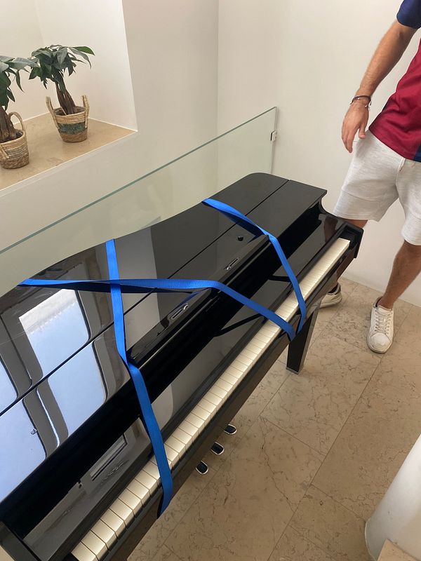 A man is moving an upright piano in a room with white walls and wooden flooring.