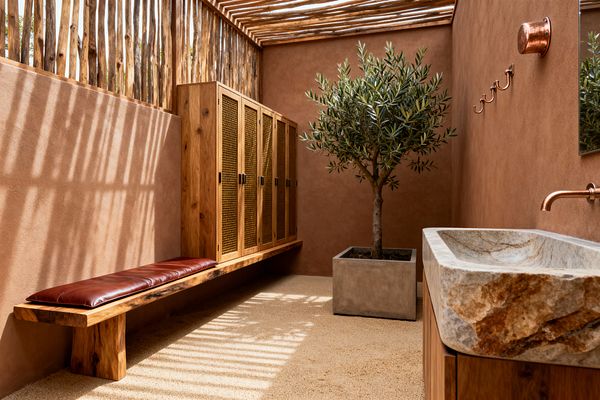 A sunlit mudroom with terracotta plaster walls and a slatted branch ceiling casting striped shadows. A long solid-wood bench with a brown leather cushion sits beside wood lockers with woven cane doors. Opposite, a large veined stone sink with a copper wall faucet and matching hooks is mounted near a mirror. An olive tree in a square concrete planter anchors the space over a sandy-textured floor.