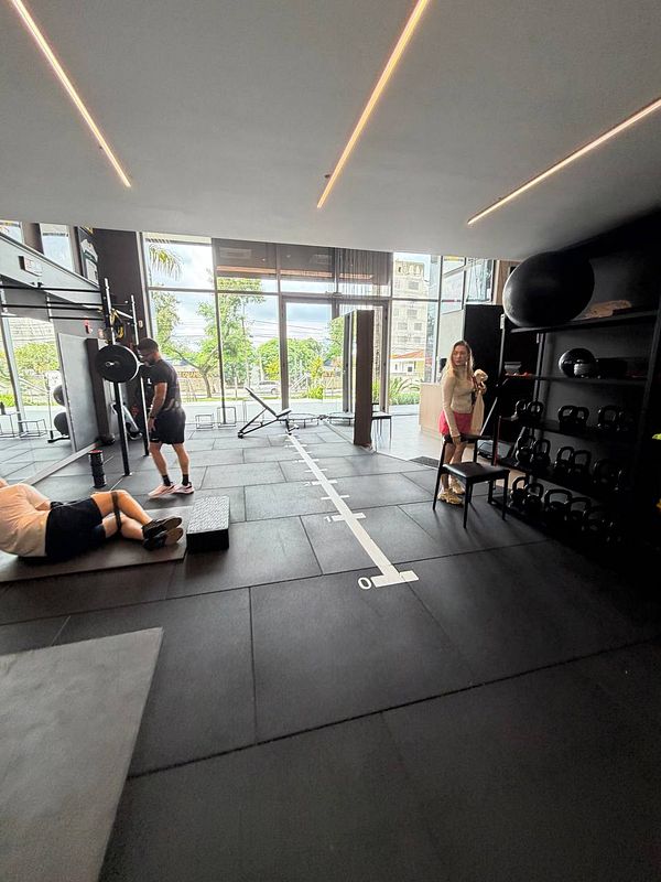 Photograph of a young woman in gym attire holding her left upper arm with her right hand, standing by a mirror in a gym setting.