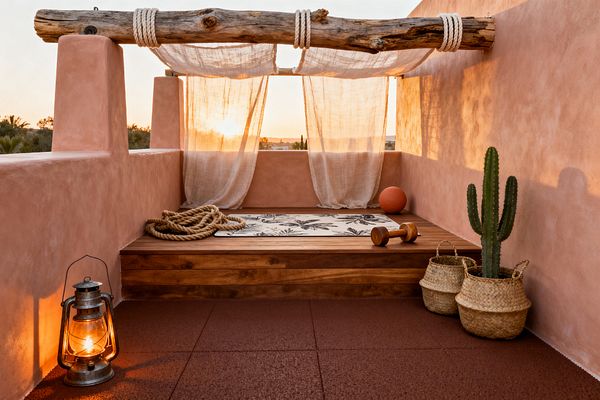 An outdoor rooftop home gym with warm terracotta stucco walls and red rubber tiles. A raised wooden platform holds a leaf-print yoga mat, a coiled battle rope, a wooden dumbbell, and an orange medicine ball. Sheer off-white curtains hang from a rope-wrapped driftwood beam, filtering the setting sun. A lit metal lantern sits on the floor, and a tall cactus stands in a woven basket beside another basket.