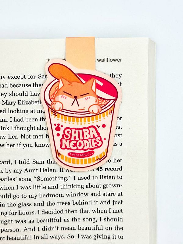 A colorful bookmark featuring a cartoon cat in a noodle cup is placed on an open book page.