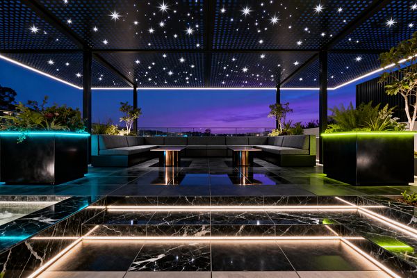 A rooftop patio at dusk shows a symmetrical U-shaped seating area under a black perforated pergola dotted with star-like LED lights and linear perimeter strips. Glossy black marble steps with warm integrated LEDs lead to charcoal modular sofas flanking two low tables with bronze cylindrical bases; black metal planters edged in neon cyan and lime hold greenery, with reflective stone flooring and a glass railing framing a purple-blue horizon.