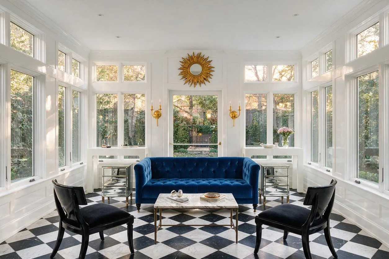 A bright sunroom with white paneled walls and many tall windows on three sides, centered around a deep blue velvet sofa. Black accent chairs, mirrored side tables, a marble coffee table, and a black-and-white diamond checkered floor create a glamorous Hollywood Regency look, with a gold sunburst mirror above the doorway.