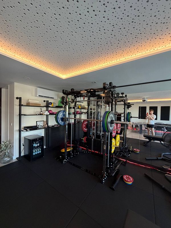 Indoor gym space with weightlifting equipment, mirror wall, and a person taking a selfie.